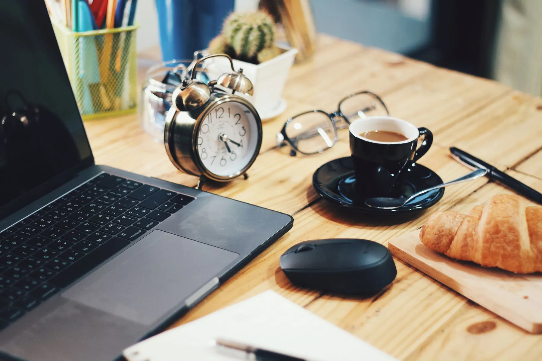 An alarm clock, a cup of coffee, a croissant and a laptop on a desk