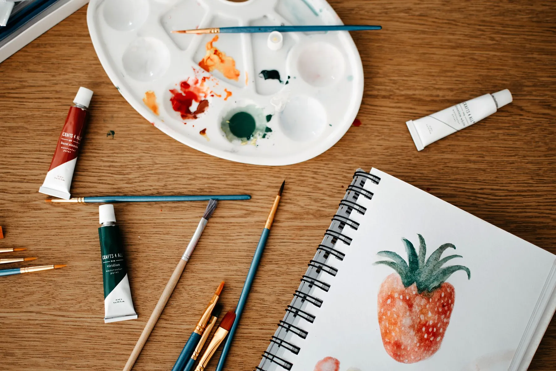 Watercolour paints, brushes and a sketchbook with a painted strawberry